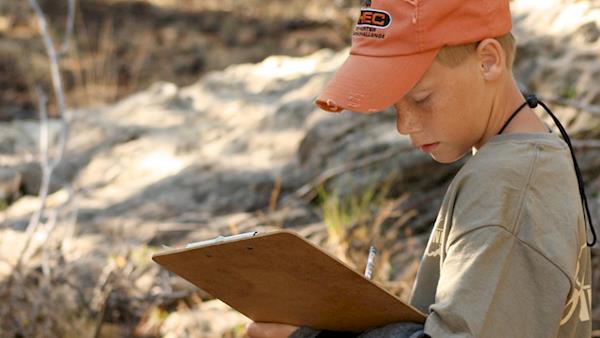 NRA Youth Hunter Education Challenge participant in an orange cap with a clip board in the wilderness.
