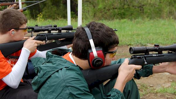 Young men wearing ear protection shooting scoped rifles at a range.